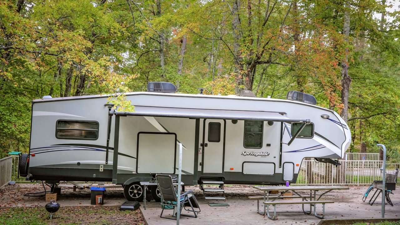 A large gray and white travel trailer sits at a campsite with an extended awning, folding chairs, and a picnic table on a concrete pad.