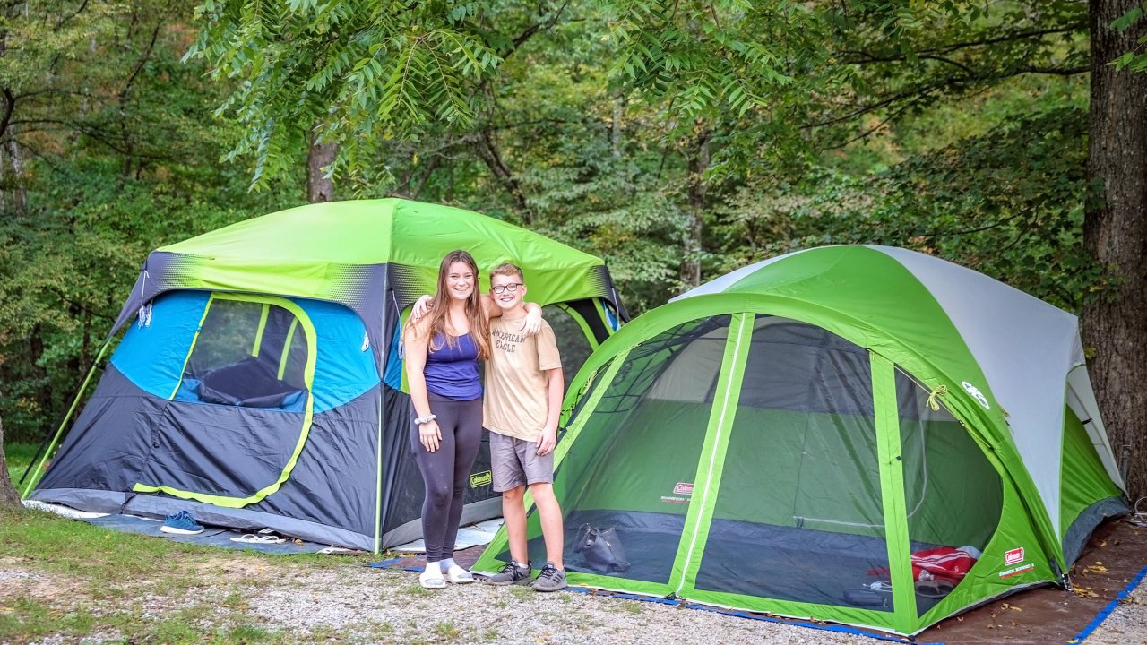 Two children stand smiling with arms around each other in front of two large green and gray camping tents set up in a wooded campsite.