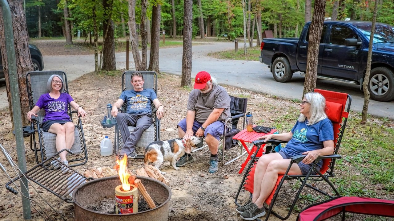Four adults sit in camping chairs around a small fire pit, chatting and relaxing while one person pets a dog in a forested campground.