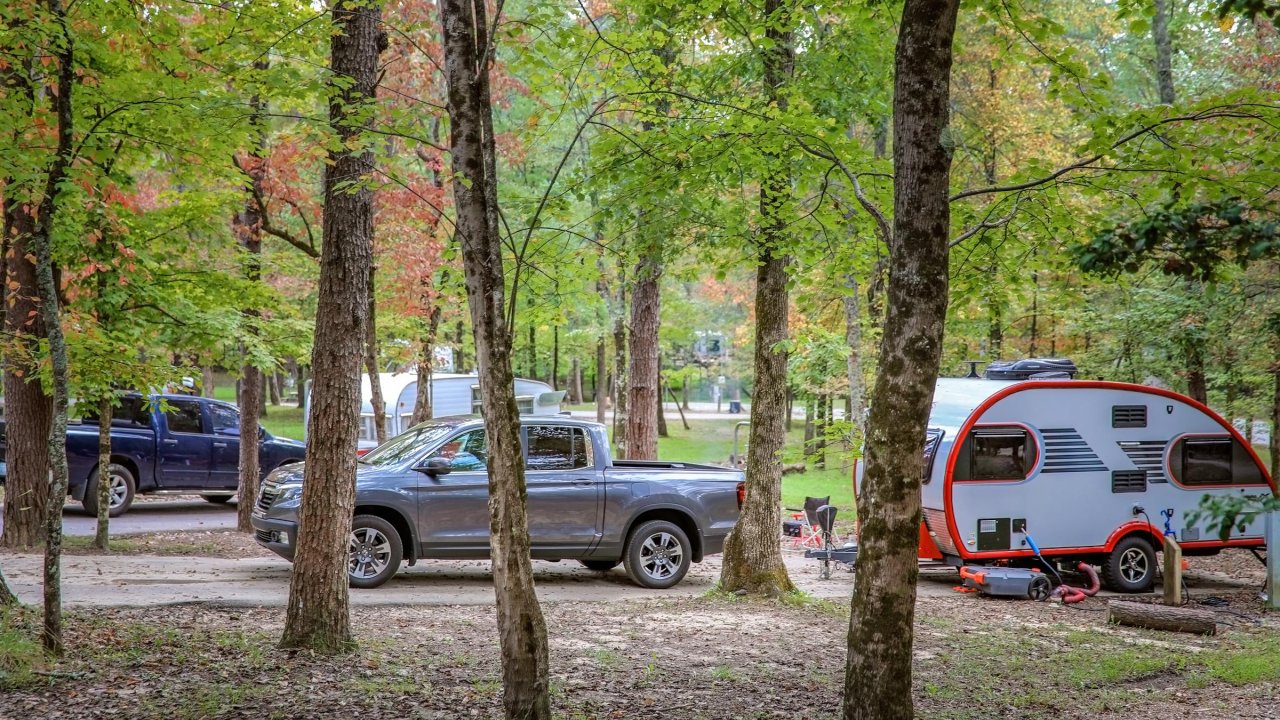 A gray pickup truck is parked next to a small red and gray camper trailer, surrounded by trees in a peaceful campsite.
