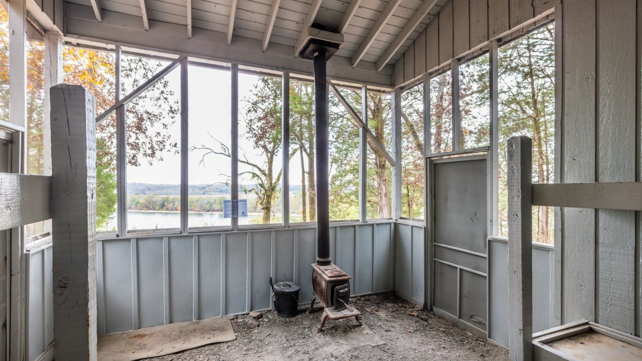 Wooden cabin interior with a stove, overlooking a lake view through large windows.