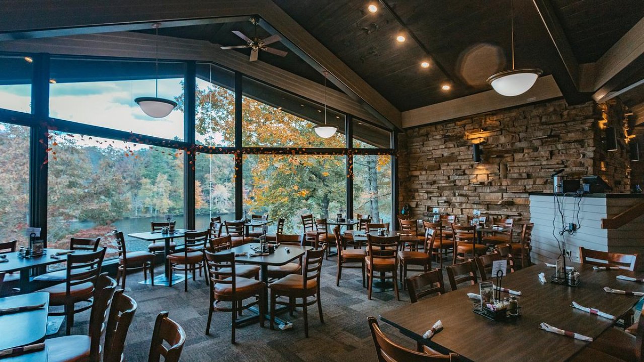 Cozy restaurant interior with large windows, wooden tables, and autumn view outside.