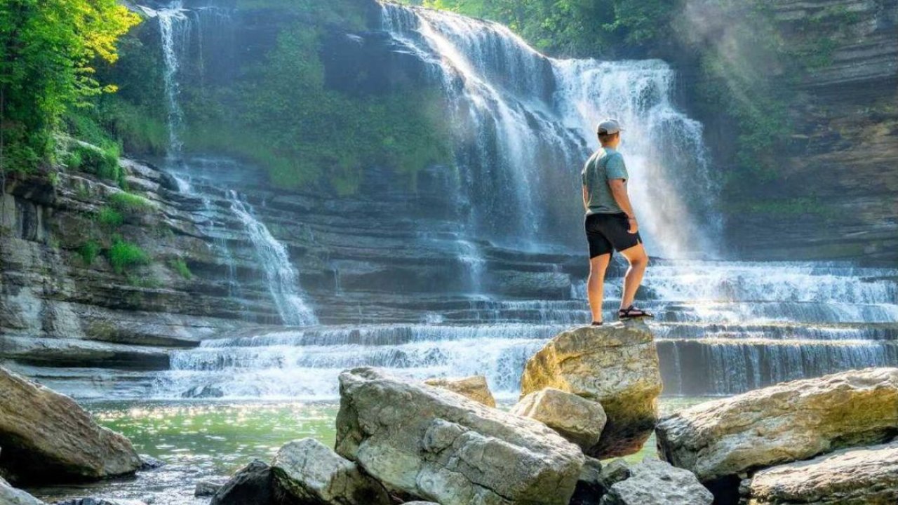 A man stands on a large rock looking out at a cascading waterfall