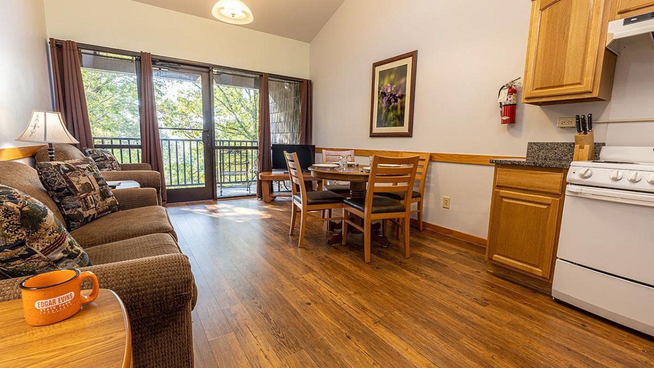 Cozy living area with sofa, table, kitchen, and balcony view. Warm lighting.