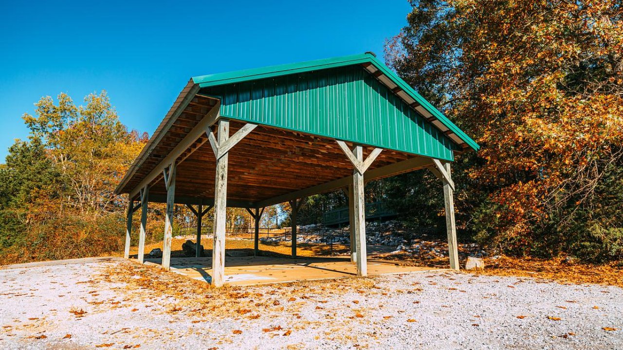 Open wooden pavilion with a green metal roof covering wheelchair-accessible parking set in a sunny, fall landscape.