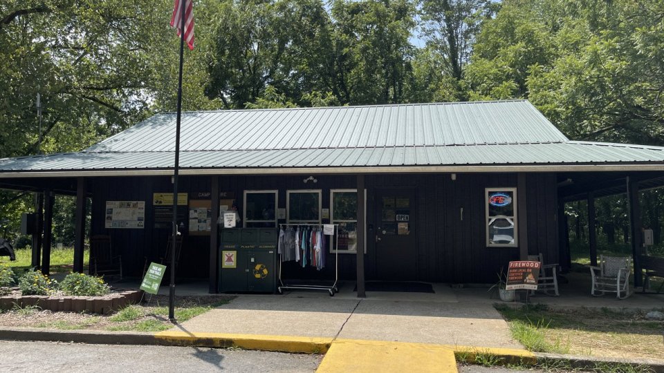 A rustic brown building with a green metal roof sits in a wooded area. Including an Icebox, and recycling bin line the porch.