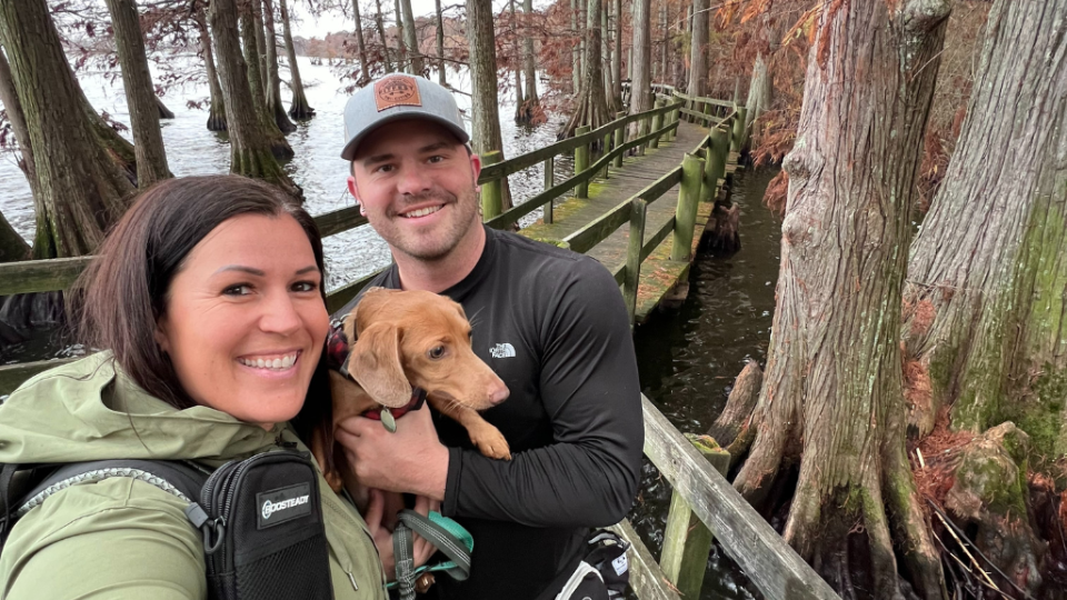 A couple poses for a selfie on a wooded boardwalk surrounded by tall trees. The man holds a small brown dog. The atmosphere is calm and outdoorsy.