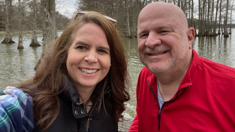 A smiling woman and man take a selfie by a lake with bare trees in the background. She wears a plaid shirt and vest; he is in a red jacket. Winter atmosphere.