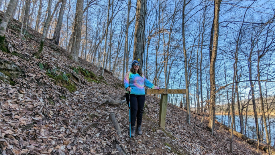 A woman wearing a tie-dye hoodie, black pants, and sunglasses stands on a forest trail beside a sign reading "Indian Trevens." The scene is peaceful with leafless trees and a lake visible in the background.