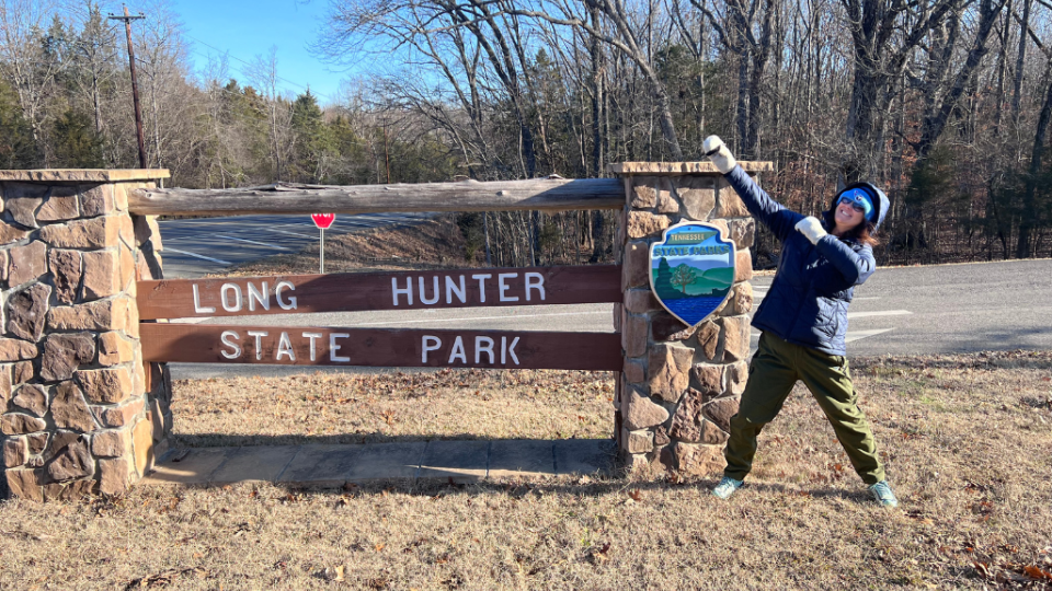 A person in winter gear playfully poses next to the Long Hunter State Park sign made of wood and stone, set against a backdrop of leafless trees and a clear road.