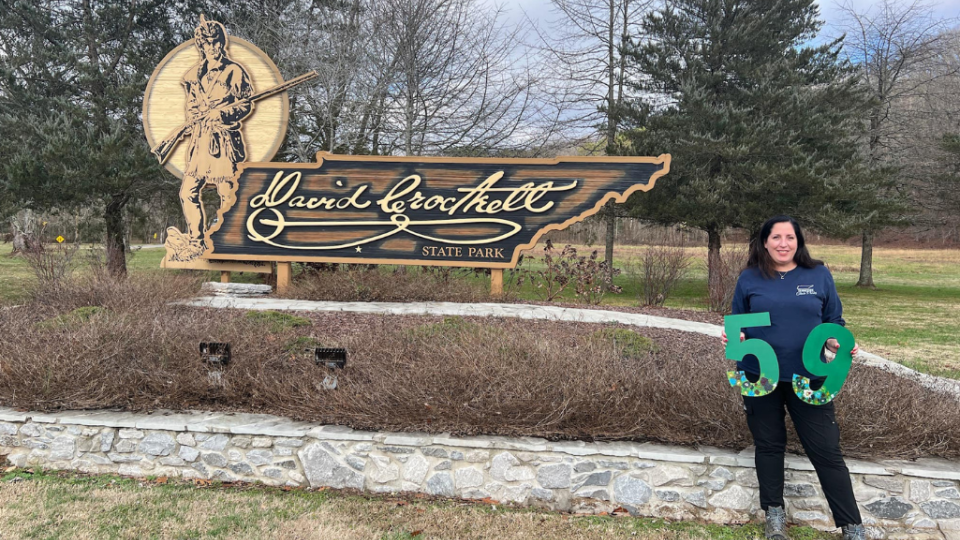 A woman stands smiling next to the David Crockett State Park sign, holding a green number "59." The park's logo features a pioneer figure. The scene is calm and welcoming.