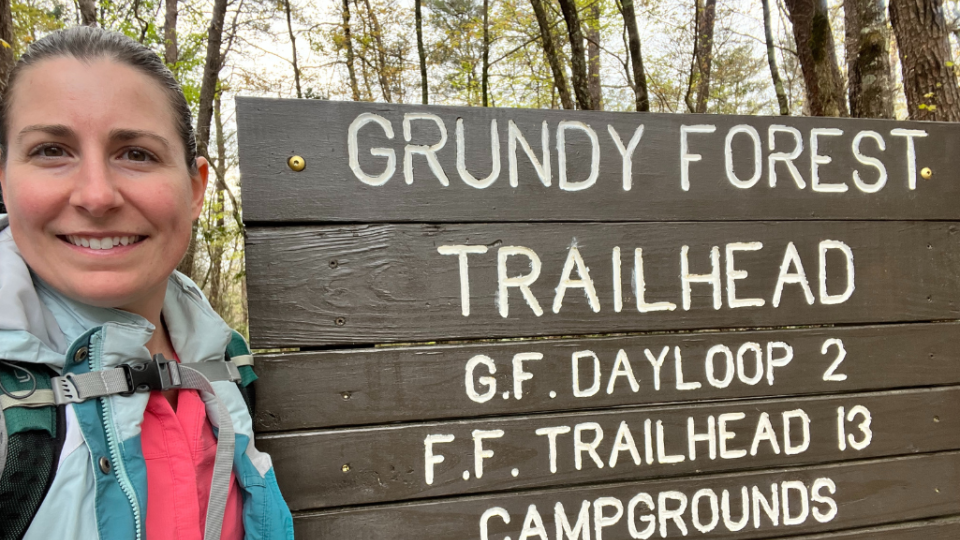 Woman smiling at Grundy Forest Trailhead sign in a wooded area. She is wearing a backpack. The atmosphere is welcoming and adventurous.