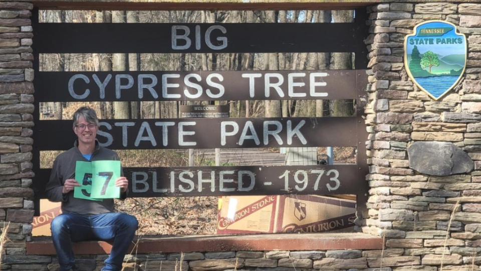 A person sits smiling next to the Big Cypress Tree State Park sign, holding a green "57" poster. The sign is set against a stone structure under a sunny sky.