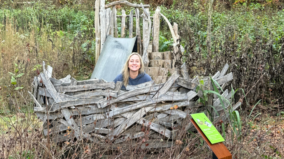 A smiling person sits behind a rustic wooden fence in a natural setting. The background shows greenery, and a small informational sign is visible nearby.