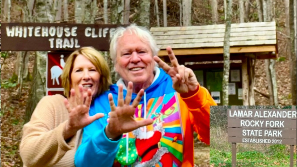 A smiling couple poses in front of the Whitehouse Cliffs Trail sign at Lamar Alexander Rocky Fork State Park. They display numbers with their fingers, wearing colorful clothing.