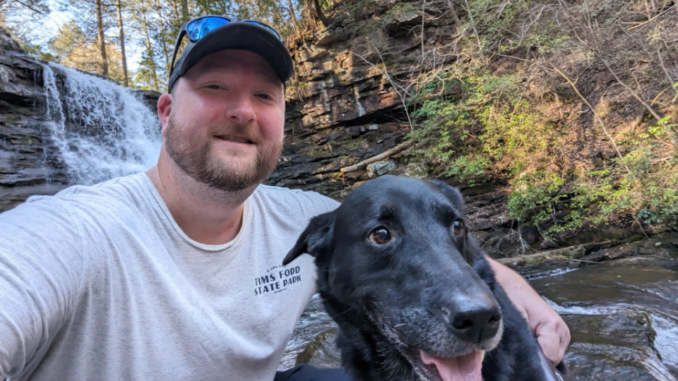 Man and black dog smiling near a waterfall in a forest. The man wears a cap and sunglasses, conveying a sense of adventure and companionship.
