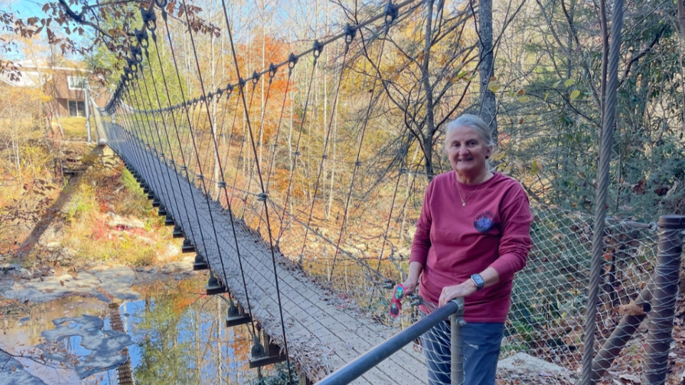 A person in a red sweater smiles while standing on a suspension bridge in a forest during autumn, with colorful leaves and a clear blue sky.
