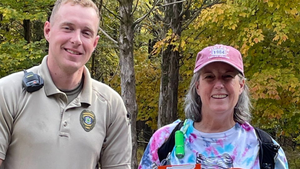 A smiling man in a uniform and a woman in a cap and tie-dye shirt pose together outdoors. Behind them, trees with autumn leaves set a warm tone.