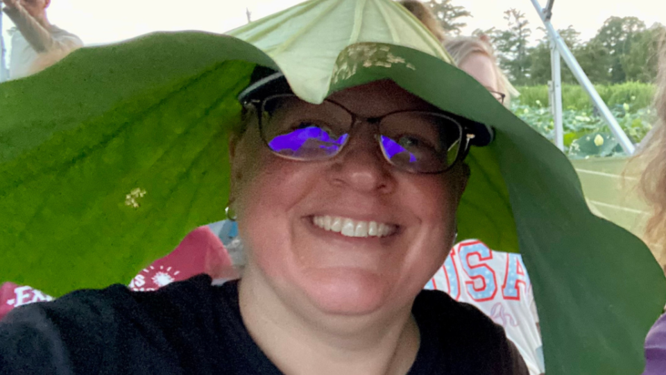 Smiling person wearing glasses and using a large green leaf as a hat. The background shows a field, other people, and a canopy structure. The mood is playful and joyful.