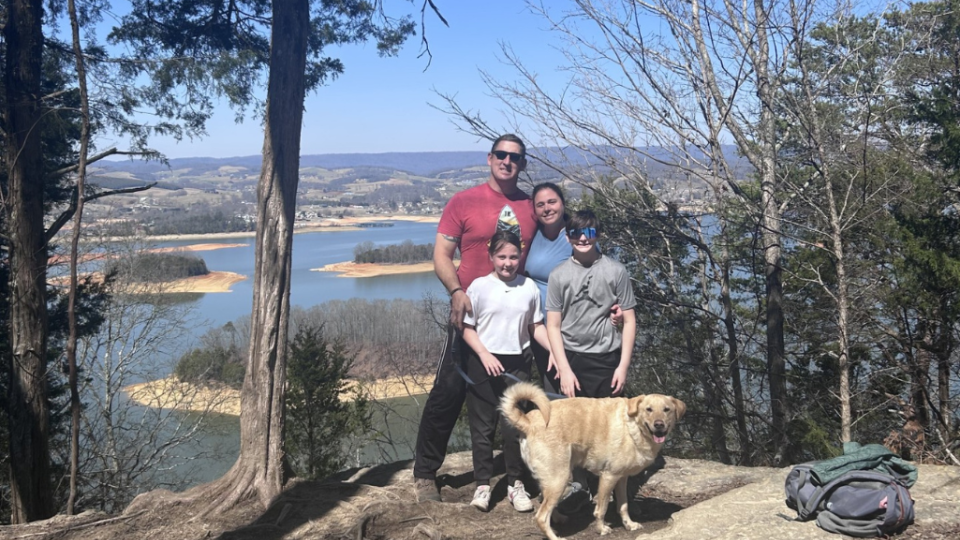 A family of four and a dog pose happily on a cliffside overlooking a scenic lake with islands. Trees frame the view, and a backpack rests on the ground.