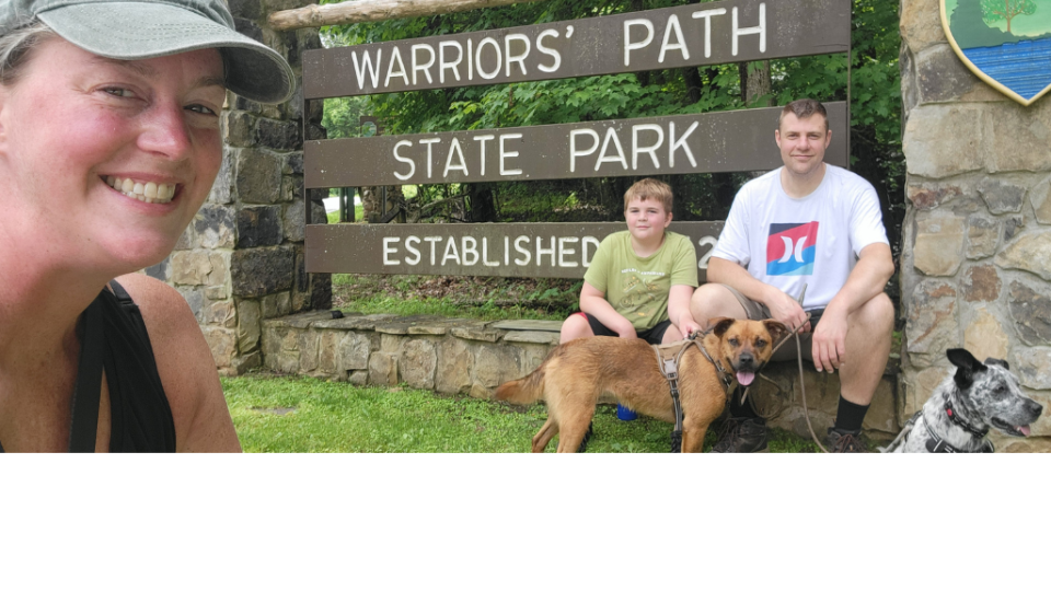 Smiling woman in cap takes selfie with two people and two dogs in front of "Warriors' Path State Park" sign. Lush greenery in the background.