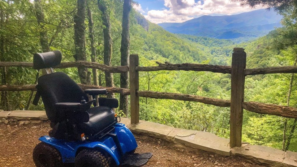 A blue all-terrain wheelchair is parked on a dirt path overlooking a lush green mountain valley.