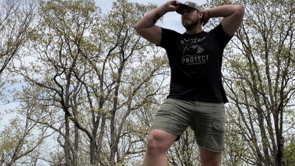 Man in a black t-shirt and khaki shorts stands confidently outdoors. He holds his cap, surrounded by tall trees under a clear sky, evoking a sense of adventure.