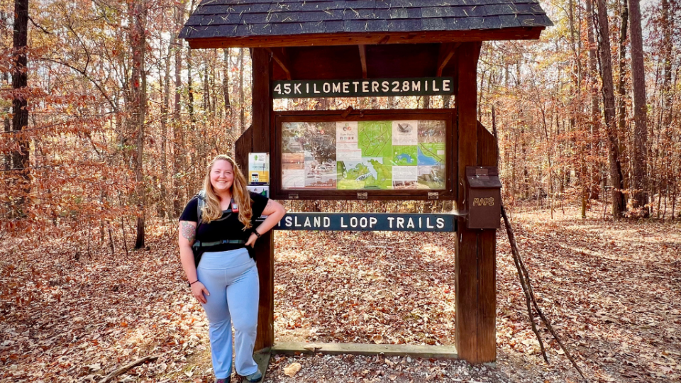 Woman smiling next to a wooden trail sign in a forest during autumn. The sign reads "4.5 kilometers 2.8 mile Island Loop Trails." Fallen leaves cover the ground.