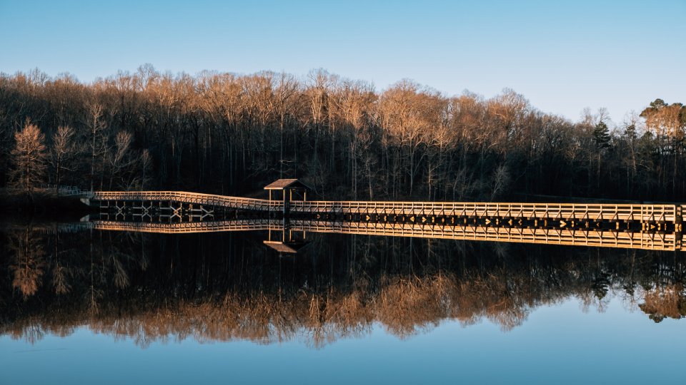 A wooden pier extends over a calm lake with a small pavilion and is mirrored in the water. Bare trees with golden light line the shore under a clear blue sky.