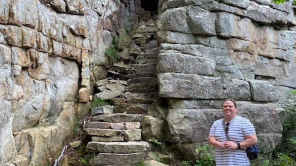 A man in a striped shirt stands smiling beside ancient stone steps leading up a rocky cliff face. The setting is rustic and suggests a sense of adventure.