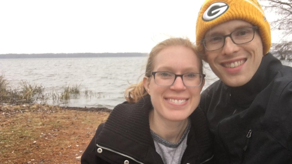 A smiling couple in jackets and glasses takes a selfie by a serene lakeside on a cloudy day. The man wears a yellow beanie with a "G" logo.