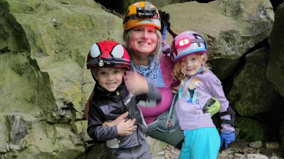 A smiling woman and two children wearing helmets with lights are posing in a rocky cave. The scene conveys adventure and joy amid the natural surroundings.