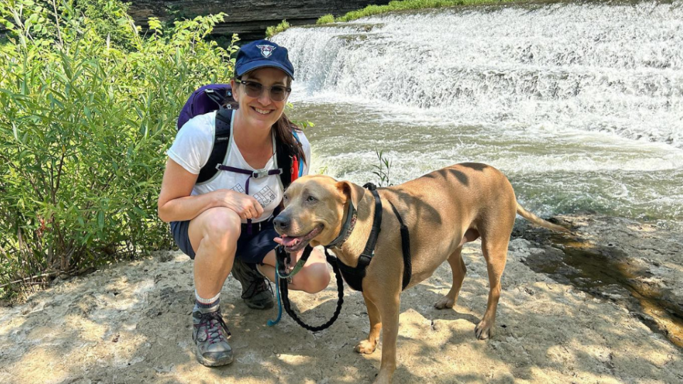 A woman wearing a cap and backpack kneels beside a happy dog near a waterfall. Sunlight filters through trees, creating a serene outdoor scene.
