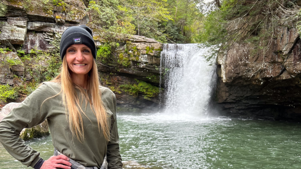 A person in a green sweater and black beanie smiles near a waterfall surrounded by lush greenery. The scene conveys a sense of adventure and serenity.