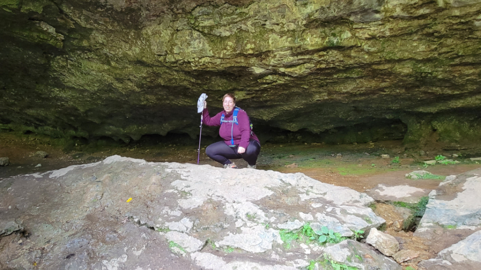 A person crouches under a rocky overhang in a cave, holding a hiking pole. The scene conveys adventure and exploration, with earthy tones.
