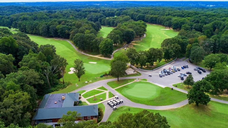 Aerial view of a lush golf course with verdant fairways, surrounded by dense trees. A clubhouse, parking lot, and multiple golf carts are visible. The scene is serene and expansive.