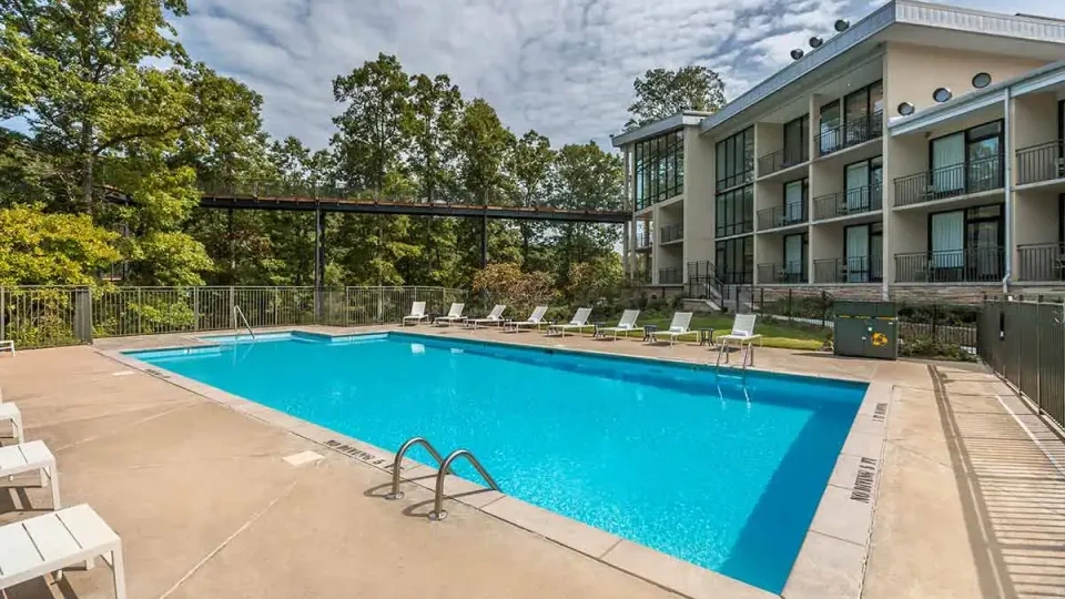 Outdoor pool with loungers, near modern building and trees under a cloudy sky.