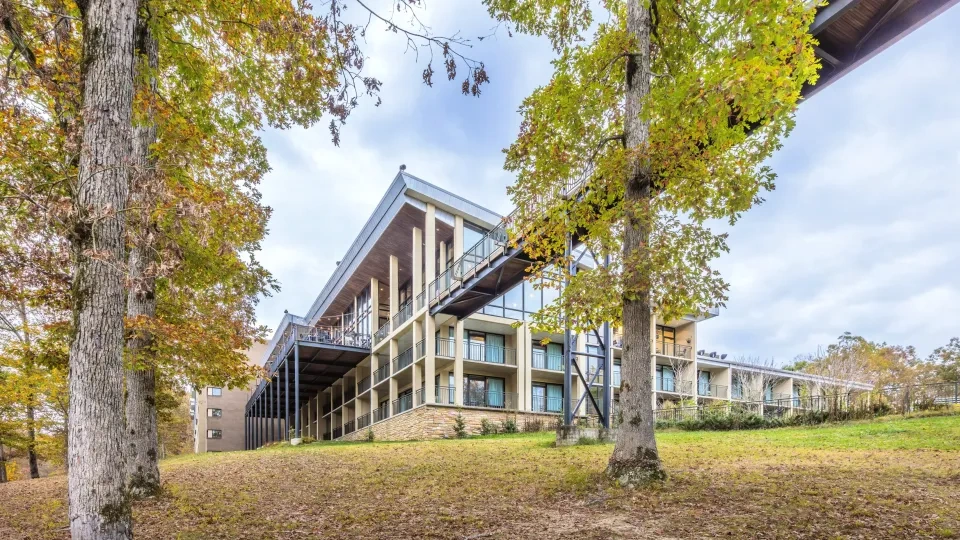 Modern building with large windows, surrounded by autumn trees and cloudy sky.
