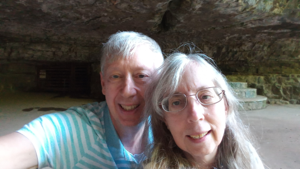 Elderly couple smiling in a dimly lit cave, with rocky walls and stacked stone steps in the background. The atmosphere feels warm and adventurous.