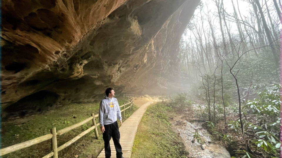 Man standing on a sidewalk under a rock formation,  looking into lush wooded area. 