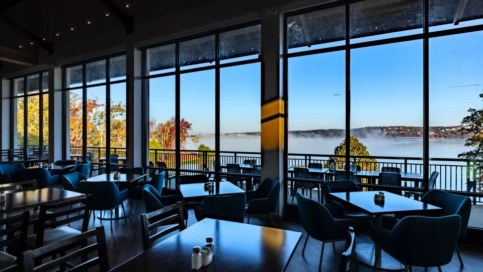 Restaurant interior with large windows overlooking a misty lake and autumn trees.