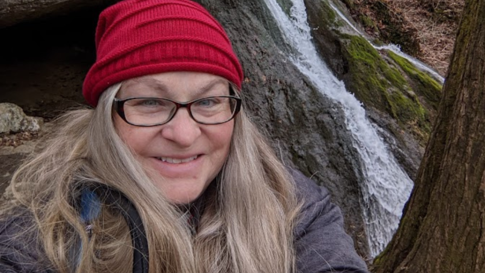 Woman in a red beanie and glasses smiling in front of a small waterfall. The scene is outdoors with mossy rocks and a tree, conveying a serene mood.