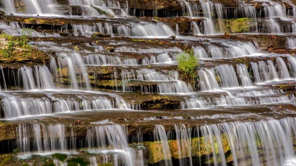 Cascading waterfall over multi-tiered rocks, water flows gently, creating a serene and tranquil atmosphere with patches of green moss and foliage.