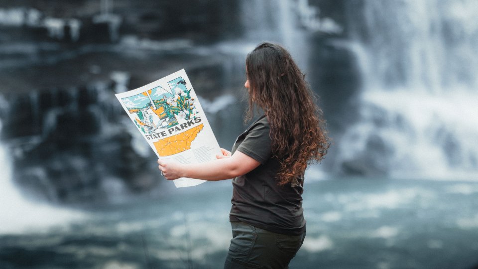 A woman stands by a waterfall, holding a colorful map titled "State Parks." Her long hair flows as she attentively looks at the map, conveying exploration.