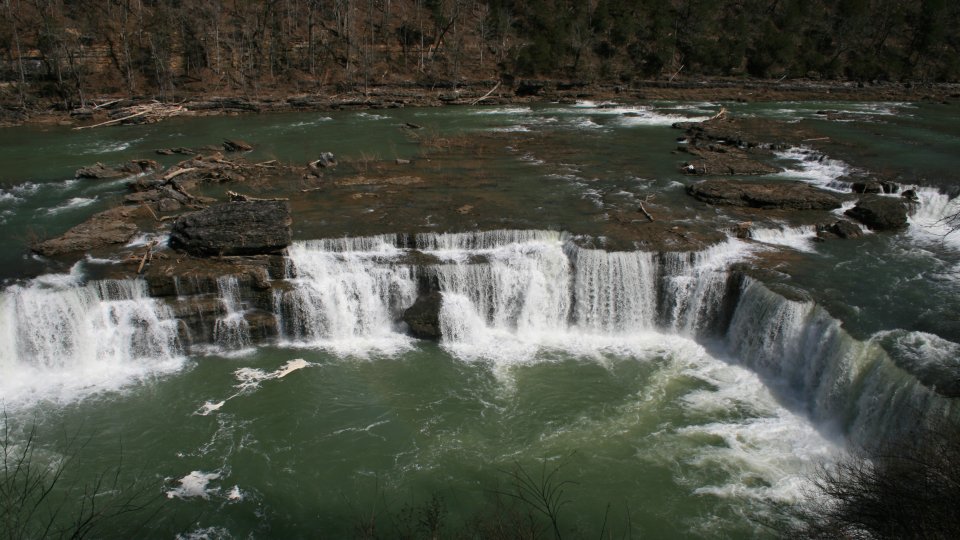 A waterfall cascades over rocky ledges into a swirling green pool, surrounded by dense forest