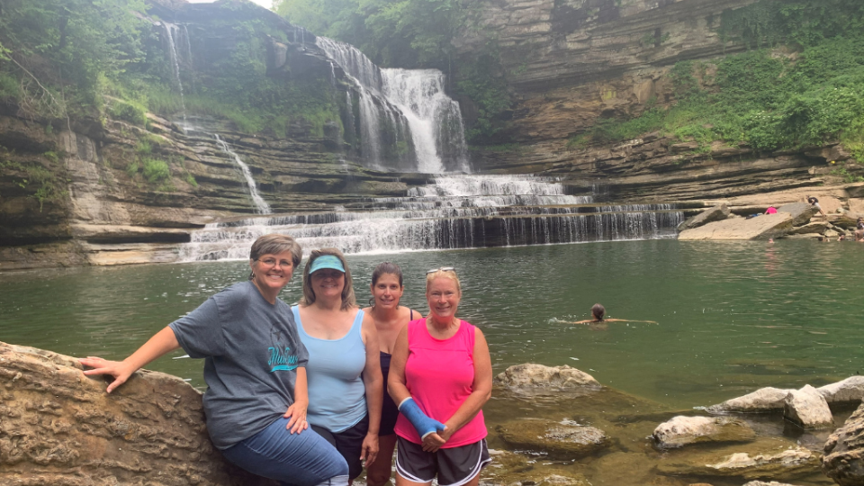 Four women smile by a serene waterfall cascading into a clear pool. Surrounded by lush greenery, the scene conveys a sense of tranquility and friendship.