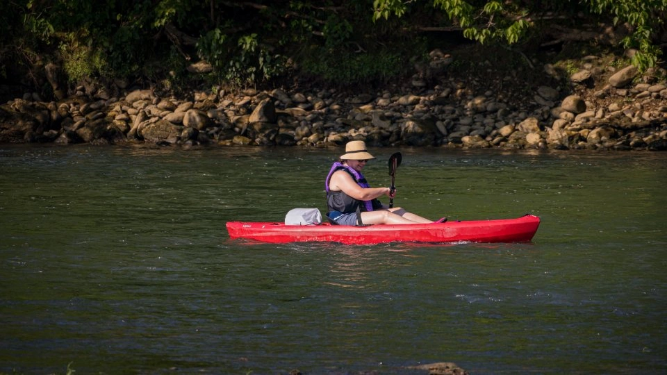 Person kayaking in a red kayak on a calm river, wearing a sun hat and life jacket. The background shows green foliage and a rocky shore, evoking tranquility.