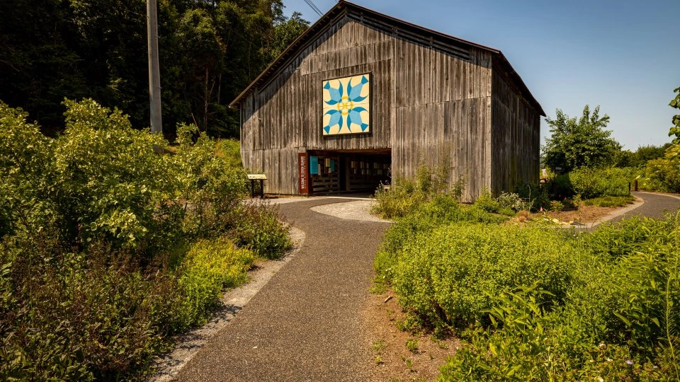 A rustic wooden barn with a colorful quilt pattern on the front, surrounded by lush greenery. A gravel path leads to its entrance under a clear blue sky.