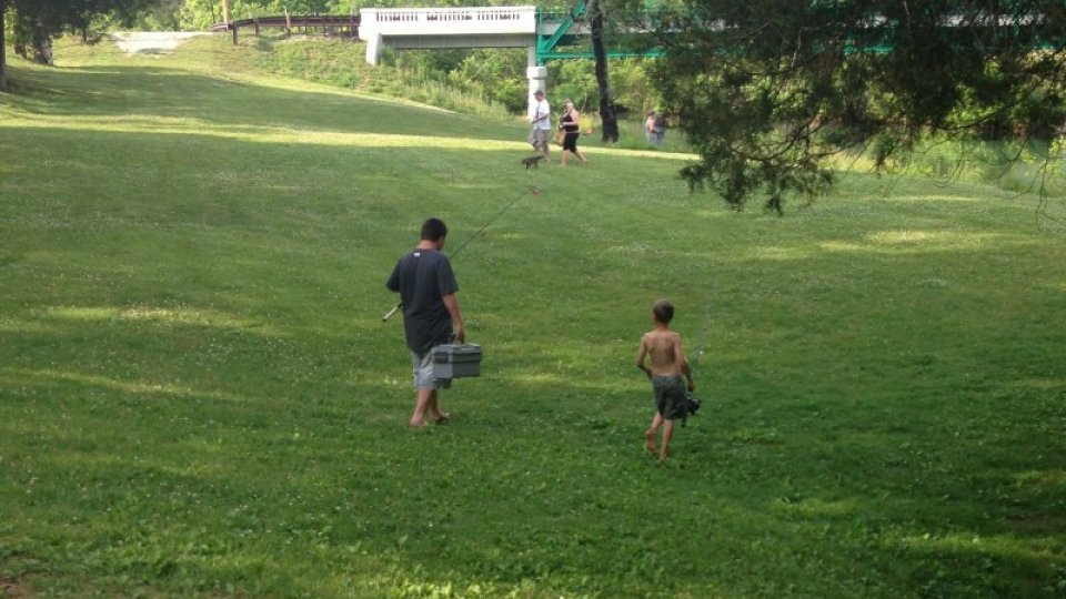A man carrying fishing gear walks with a shirtless boy across a lush, green park. People in the background enjoy the sunny day near a bridge.