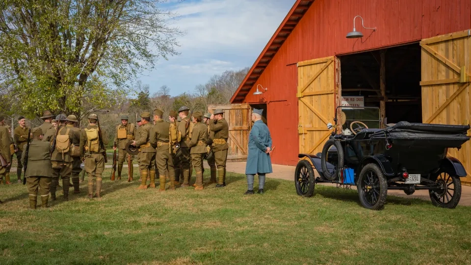 A group of people in vintage military uniforms stand outside a red barn with wooden doors. A vintage black car is parked nearby on a grassy field. The atmosphere is historical and reflective.